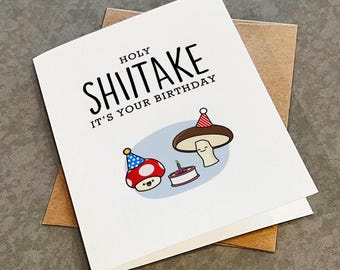 May include: A white greeting card with the text "HOLY SHITake IT'S YOUR BIRTHDAY" in black. The card features a cartoon illustration of two mushrooms wearing party hats and a birthday cake with a single candle.