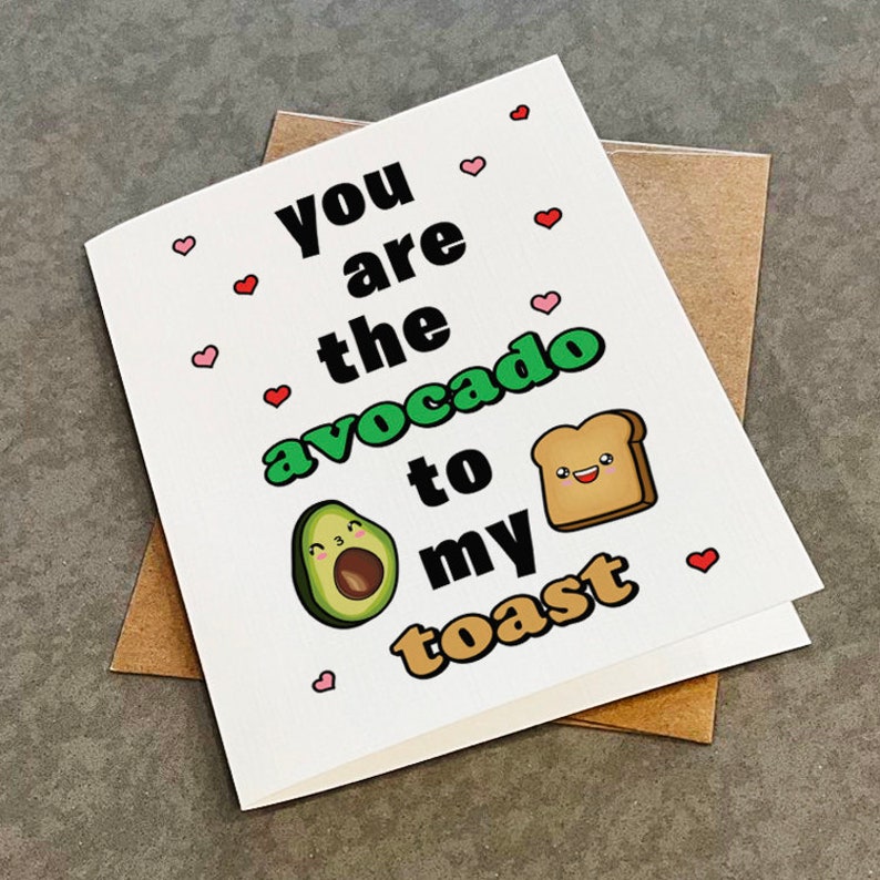 Pu&ograve; includere: Biglietto di auguri bianco con la scritta "You are the avocado to my toast". Il biglietto presenta illustrazioni di un avocado e una fetta di pane tostato, con piccoli cuori rossi e rosa.