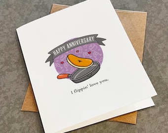 May include: A white greeting card featuring a cartoon illustration of a pancake flipping in a frying pan. Above the pan is a grey banner with the text HAPPY ANNIVERSARY. Below the illustration is the text I flippin' love you.