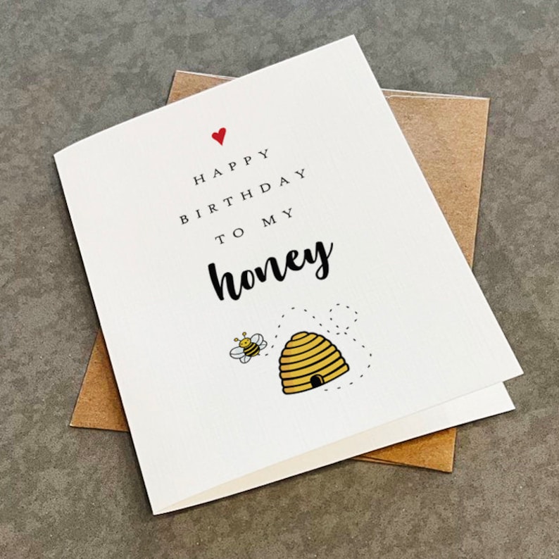 May include: A white card with a red heart, black text that says "Happy Birthday to my honey" and a cartoon bee flying towards a beehive.