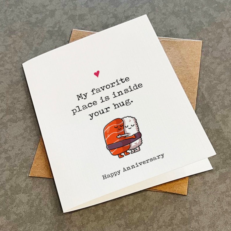 May include: A white card with a pink heart and the text "My favourite place is inside your hug. Happy Anniversary."  A cartoon illustration of two pieces of sushi hugging each other.