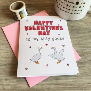 May include: A white card with pink hearts and the text "Happy Valentine's Day to my silly goose" in red letters. Two white geese are illustrated on the card, one facing left and the other facing right.