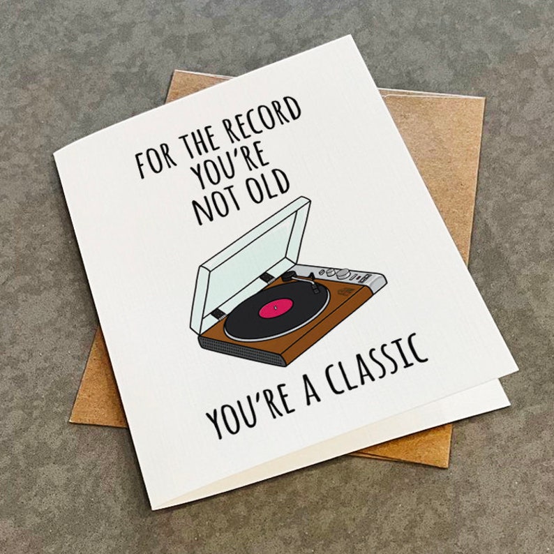 May include: A white card with black text that reads "For the record you're not old. You're a classic."  A black and white illustration of a record player with a brown record on top is in the center of the card.