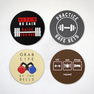 May include: Four round coasters with motivational fitness quotes and images. The first coaster is black with red text that says "No Pain No Gain Raise The Bar" and a barbell image. The second coaster is grey with white text that says "Practice Safe Sets" and a barbell image. The third coaster is beige with red and blue kettlebells and text that says "Grab Life By The Bells". The fourth coaster is brown with white text that says "Lift Eat Sleep Repeat" and images of a barbell, a plate with a fork and knife, and a bed.