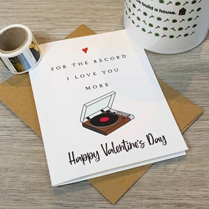 May include: A white card with a red heart and a black and white illustration of a record player. The text on the card reads "For the record I love you more Happy Valentine's Day".