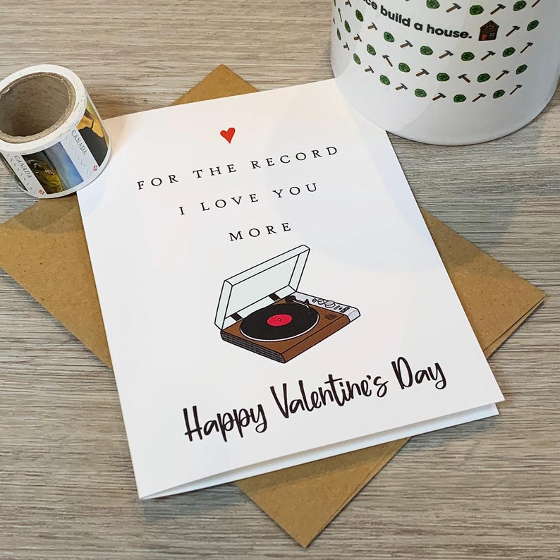 May include: A white card with a red heart and a black and white illustration of a record player. The text on the card reads "For the record I love you more Happy Valentine's Day".