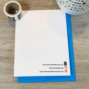 May include: A white card with blue text on the bottom. The text reads "www.lettucebuildahouse.com", "@lettucebuildahouse", and "www.lettucebuildahouse.etsy.com". The card is on top of a blue envelope.