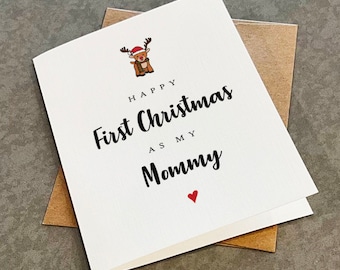 May include: A white Christmas card with the message "Happy First Christmas as my Mommy" in black script. A cartoon reindeer wearing a Santa hat and scarf is at the top. A small red heart is at the bottom.