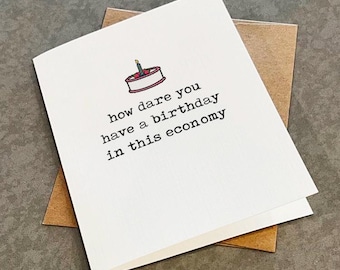 May include: A white greeting card featuring a small illustration of a birthday cake with a single lit candle at the top. Below the cake, the text reads: how dare you have a birthday in this economy. The card is placed on top of a brown envelope.