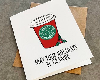 May include: A white greeting card featuring a red coffee cup illustration with a green circular logo that says Happy Holidays and a small Christmas tree icon. Below the cup, the text reads May Your Holidays Be Grande.