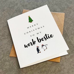 May include: A white Christmas card with the message "Merry Christmas to my work bestie." The card features a green Christmas tree and two cartoon penguins wearing Santa hats. The card is inside a brown envelope.
