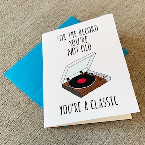 May include: A white card with a blue envelope. The card has a black and white illustration of a record player with a red record. The text on the card reads "For the record you're not old you're a classic".