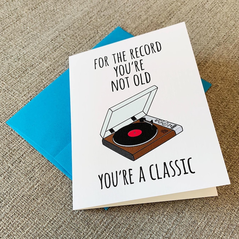 May include: A white card with a blue envelope. The card has a black and white illustration of a record player with a red record. The text on the card reads "For the record you're not old you're a classic".