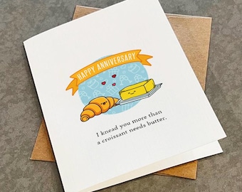May include: A white greeting card with a brown envelope. The card features a cartoon illustration of a croissant and butter with the text "Happy Anniversary" and "I knead you more than a croissant needs butter."