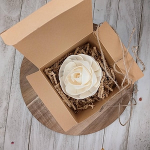 May include: A cream-colored, wooden flower rests inside an open, brown cardboard box. The box is lined with brown paper shreds. The box sits on a wooden surface. Jute twine is wrapped around the box.