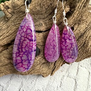 May include: A jewelry set consisting of a teardrop-shaped pendant on a black cord and two matching teardrop-shaped earrings. Each piece features a polished purple stone with a dark, web-like crackle pattern, secured with silver-toned wire loops.