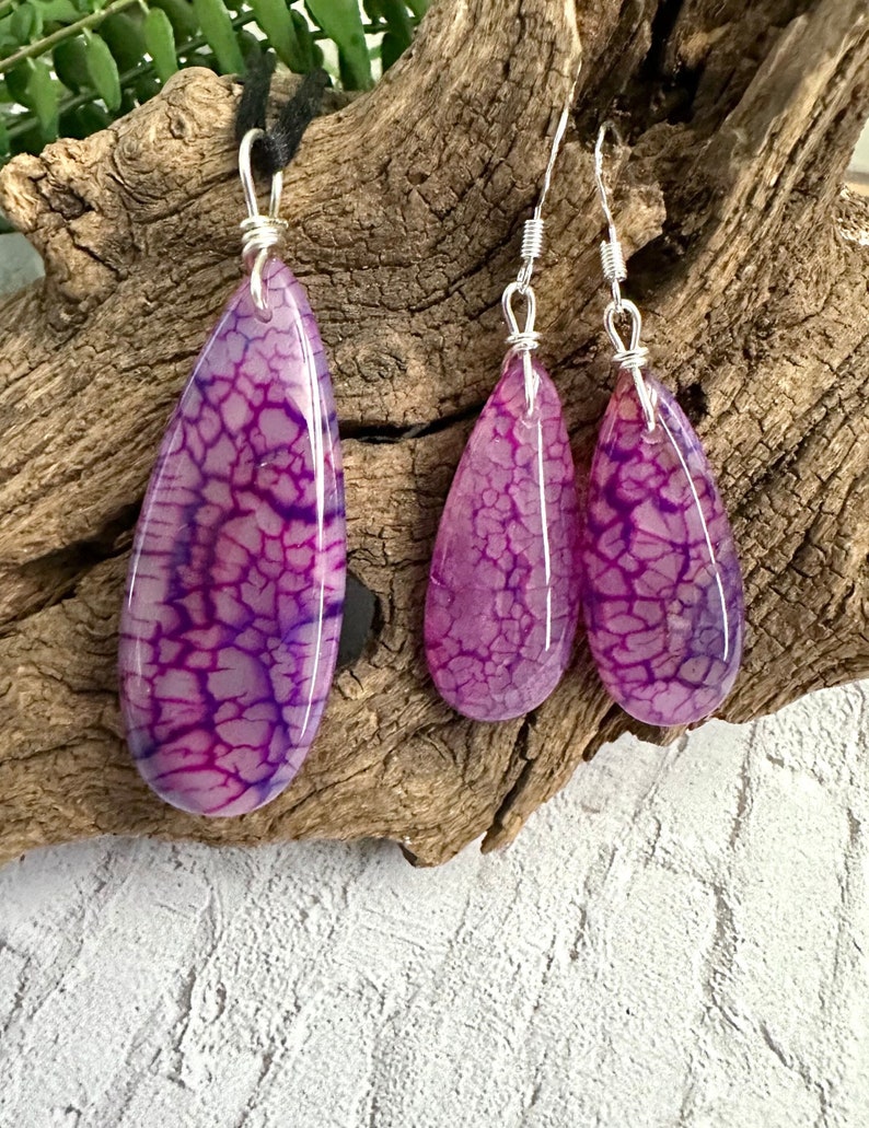 May include: A jewelry set consisting of a teardrop-shaped pendant on a black cord and two matching teardrop-shaped earrings. Each piece features a polished purple stone with a dark, web-like crackle pattern, secured with silver-toned wire loops.