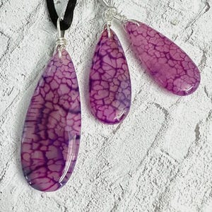May include: A jewelry set consisting of a pendant on a black cord and two matching drop earrings. Each piece features a smooth, teardrop-shaped purple stone with a dark, web-like crackle pattern, secured with silver-toned wire wrapping.