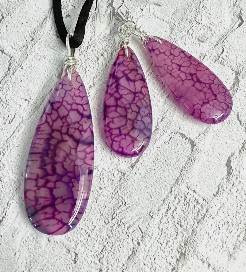 May include: A jewelry set consisting of a pendant on a black cord and two matching drop earrings. Each piece features a smooth, teardrop-shaped purple stone with a dark, web-like crackle pattern, secured with silver-toned wire wrapping.