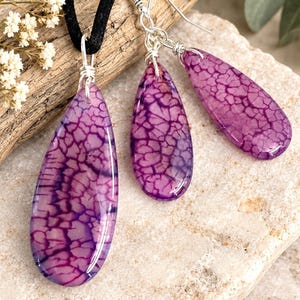 May include: A jewelry set consisting of a pendant on a black cord and two matching drop earrings. Each piece features a teardrop-shaped purple stone with a dark, crackled web pattern, secured with silver-toned wire wraps and earring hooks.