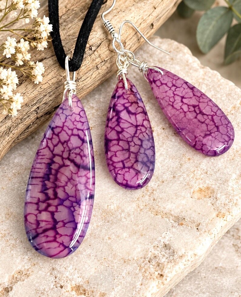 May include: A jewelry set consisting of a pendant on a black cord and two matching drop earrings. Each piece features a teardrop-shaped purple stone with a dark, crackled web pattern, secured with silver-toned wire wraps and earring hooks.