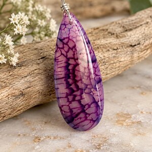 May include: A teardrop-shaped pendant featuring a purple and white crackled stone pattern. The polished stone is suspended from a silver-toned metal bail attached to a black cord necklace, showcasing a distinct web-like texture throughout the surface.
