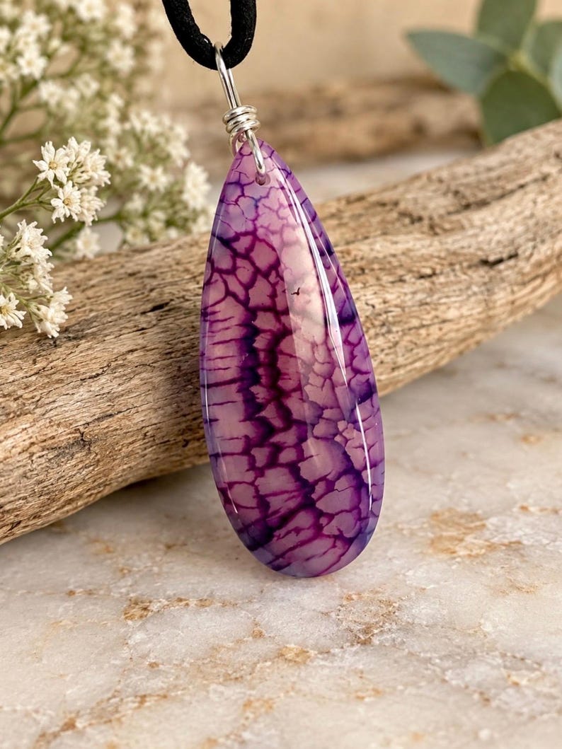 May include: A teardrop-shaped pendant featuring a purple and white crackled stone pattern. The polished stone is suspended from a silver-toned metal bail attached to a black cord necklace, showcasing a distinct web-like texture throughout the surface.
