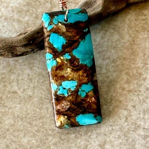 May include: Rectangular pendant with a striking pattern of turquoise blue and brown hues, with gold accents. The pendant has a copper-coloured wire loop for hanging. The stone is approximately 3.8 cm long.