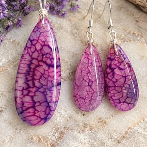 May include: A jewelry set consisting of a teardrop-shaped pendant on a black cord and matching teardrop earrings. Each piece features a polished, vibrant magenta and purple crackled stone pattern with silver-toned wire wrapping and hooks.