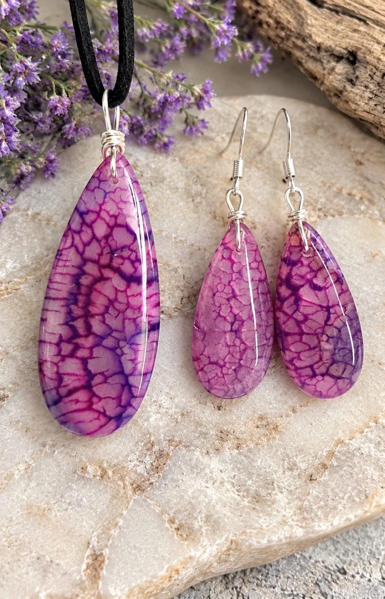May include: A jewelry set consisting of a teardrop-shaped pendant on a black cord and matching teardrop earrings. Each piece features a polished, vibrant magenta and purple crackled stone pattern with silver-toned wire wrapping and hooks.