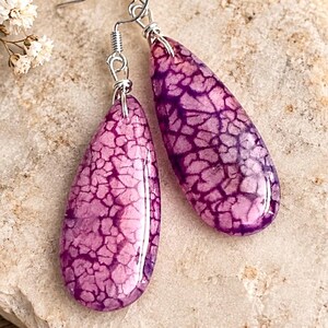 May include: A pair of teardrop-shaped earrings featuring a vibrant purple crackle pattern on a smooth, polished surface. The earrings are suspended from silver-toned metal fishhook wires, showcasing a distinct web-like texture across the purple stone.