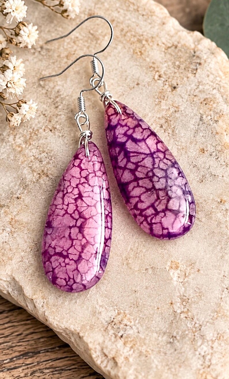 May include: A pair of teardrop-shaped earrings featuring a vibrant purple crackle pattern on a smooth, polished surface. The earrings are suspended from silver-toned metal fishhook wires, showcasing a distinct web-like texture across the purple stone.