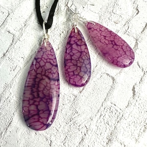 May include: A jewelry set consisting of a pendant on a black cord and two matching drop earrings. Each piece features a smooth, teardrop-shaped purple stone with a dark, web-like crackle pattern, secured with silver-toned wire wrapping.