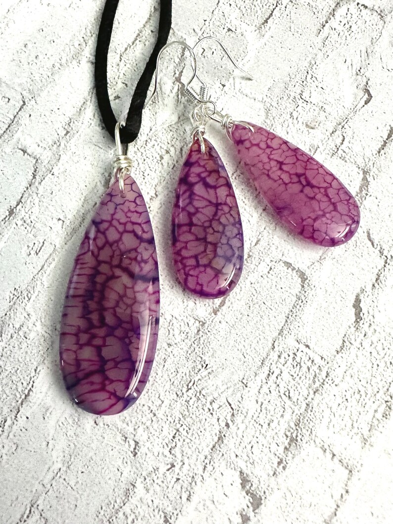 May include: A jewelry set consisting of a pendant on a black cord and two matching drop earrings. Each piece features a smooth, teardrop-shaped purple stone with a dark, web-like crackle pattern, secured with silver-toned wire wrapping.