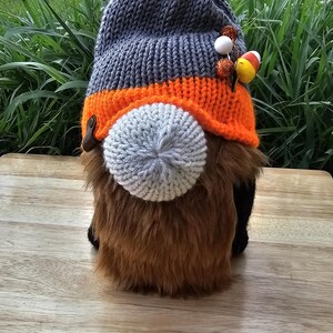 May include: A gray and orange knitted gnome hat with a yellow pom-pom and orange and white candy corn accents. The gnome is wearing a gray and orange knitted hat with a yellow pom-pom and orange and white candy corn accents. The gnome is sitting on a wooden surface.