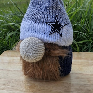 May include: A gray and white knitted gnome hat with a blue pom pom and a black star. The gnome has a brown beard and a white knitted nose.