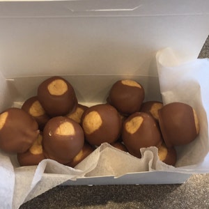 May include: A white box filled with chocolate-covered peanut butter balls. The candies are brown with a tan circle on top, resembling buckeye nuts. The box is lined with white paper, and the candies are arranged inside.