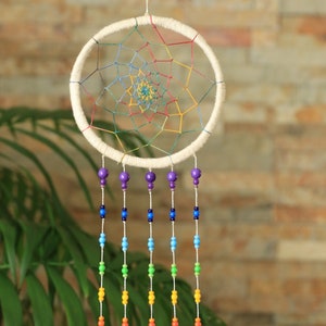May include: A handcrafted dreamcatcher with a cream-colored circular frame and a colorful web design. Strands of beads in rainbow colors hang below, leading to bamboo tubes. The dreamcatcher is suspended by a white string.