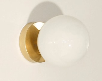 Könnte beinhalten: Moderne Wandleuchte mit einer runden, weißen Glaskugel und einem goldfarbenen, halbkreisförmigen Sockel. Die Leuchte ist an einer weißen Wand montiert und wirft einen Schatten. Das Design ist minimalistisch und modern.