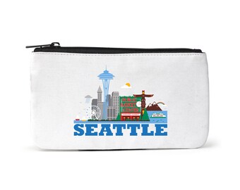 Puede incluir: Una bolsa de lona blanca con cremallera que muestra una ilustración colorida del horizonte de Seattle, incluyendo la Space Needle, el mercado Pike Place y el icónico tótem. El texto "SEATTLE" está impreso en azul debajo de la ilustración.