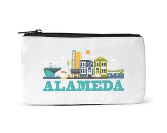 Puede incluir: Una bolsa de lona blanca con cremallera que muestra una ilustración colorida del horizonte de Alameda, que incluye un barco, edificios y un puente. El texto "ALAMEDA" está impreso en color turquesa debajo de la ilustración.