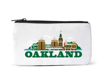 Puede incluir: Una bolsa de lona blanca con cremallera con una ilustración verde y blanca del horizonte de Oakland. El texto "OAKLAND" está impreso en letras verdes debajo del horizonte.