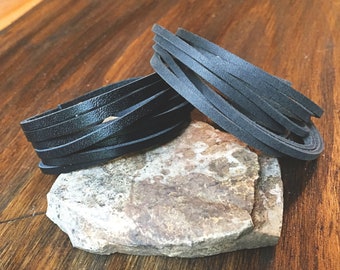 May include: Two black leather bracelets, one with a braided design and the other with a layered design, are displayed on a rough, gray rock.