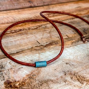 Puede incluir: Un collar artesanal con un cordón de cuero marrón rojizo y una cuenta cilíndrica turquesa con un diseño en espiral. El collar tiene un cierre de cobre. La joya se exhibe sobre una superficie de madera.