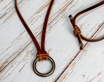 May include: A brown leather necklace with a brass ring pendant. The necklace has a simple design and is tied with a knot at the end.