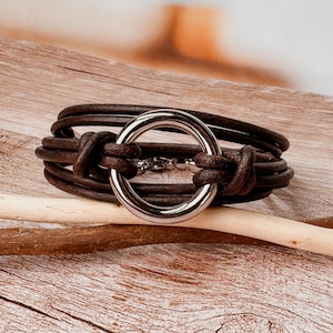 May include: A dark brown leather cord bracelet featuring multiple strands wrapped around a central polished silver-tone metal ring. The leather cords are secured with knots on either side of the circular metal component, creating a layered design.