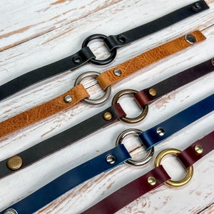 May include: Five leather bracelets with different coloured leather and metal rings. The bracelets are arranged in a row on a white wooden surface.