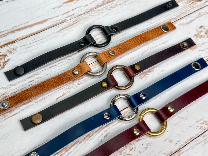 May include: Five leather bracelets with different coloured leather and metal rings. The bracelets are arranged in a row on a white wooden surface.