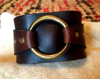 May include: A brown leather cuff bracelet with a large brass ring. The bracelet has brown leather straps and brass rivets.