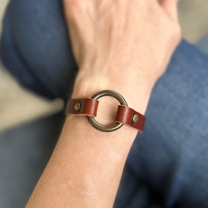 May include: A brown leather bracelet with a large, circular, antique brass-coloured metal ring.